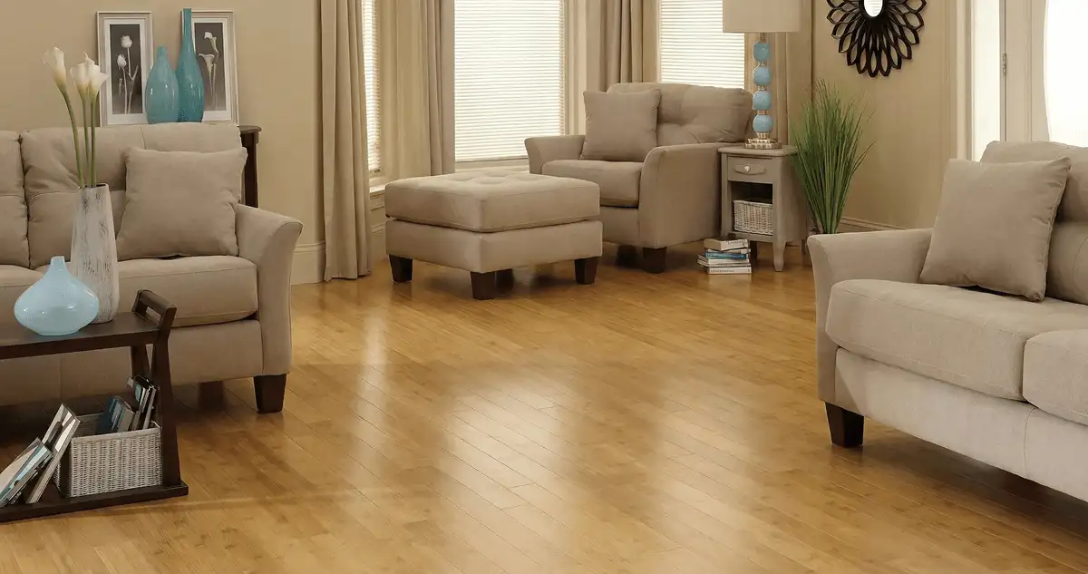 A living room with professionally installed bamboo flooring, beige sofas, a matching ottoman, a side table with a blue lamp, vases, framed pictures, and large windows with blinds and curtains in Kansas City, KS